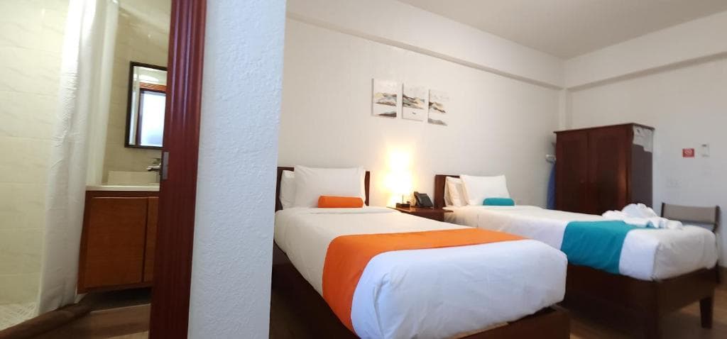 A hotel room featuring two beds with orange and teal accents and a bathroom area visible in the background.