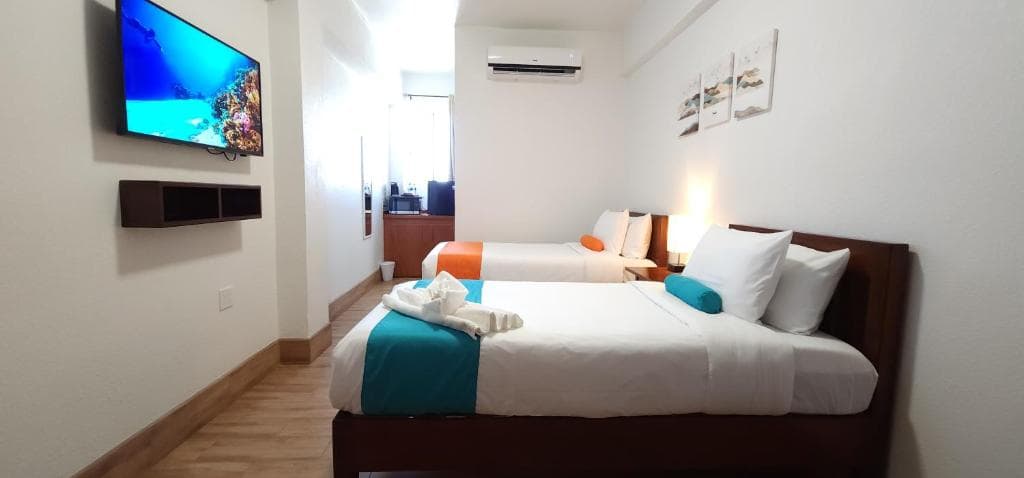 A bright hotel room features two beds with colorful accents and a wall-mounted TV.