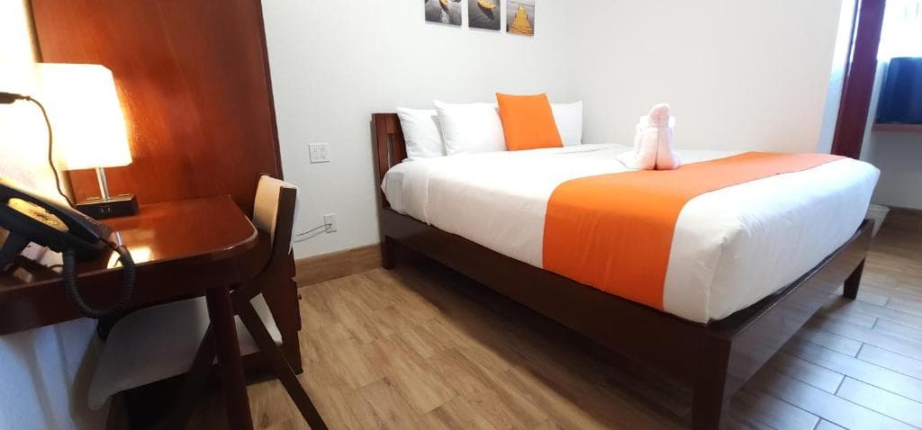 A neatly arranged hotel room featuring a bed with an orange accent, a wooden desk, and decorative wall art.