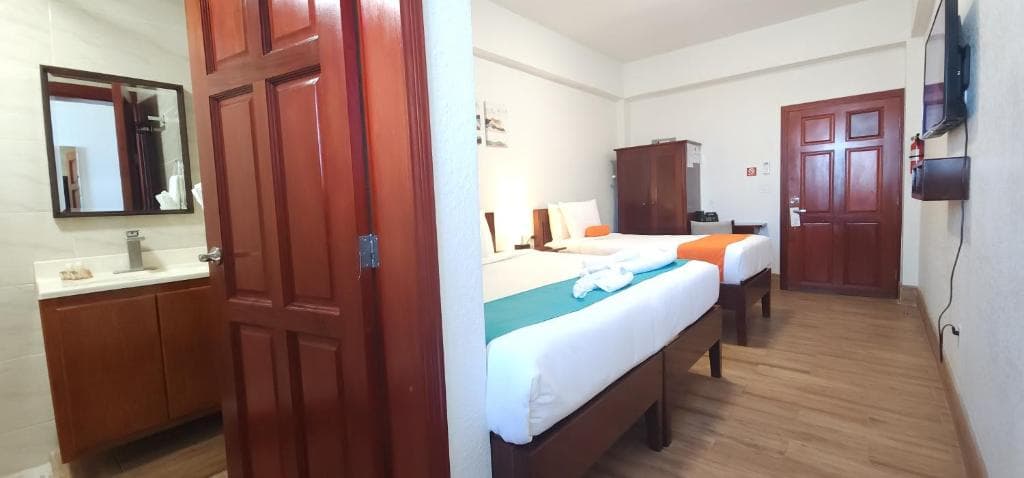 A brightly lit hotel room featuring two beds, a wooden door, and a bathroom area.