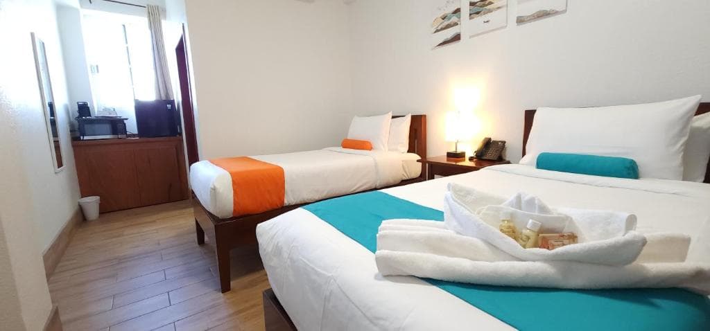 A well-lit hotel room featuring two beds with colorful accents and a small kitchenette area.