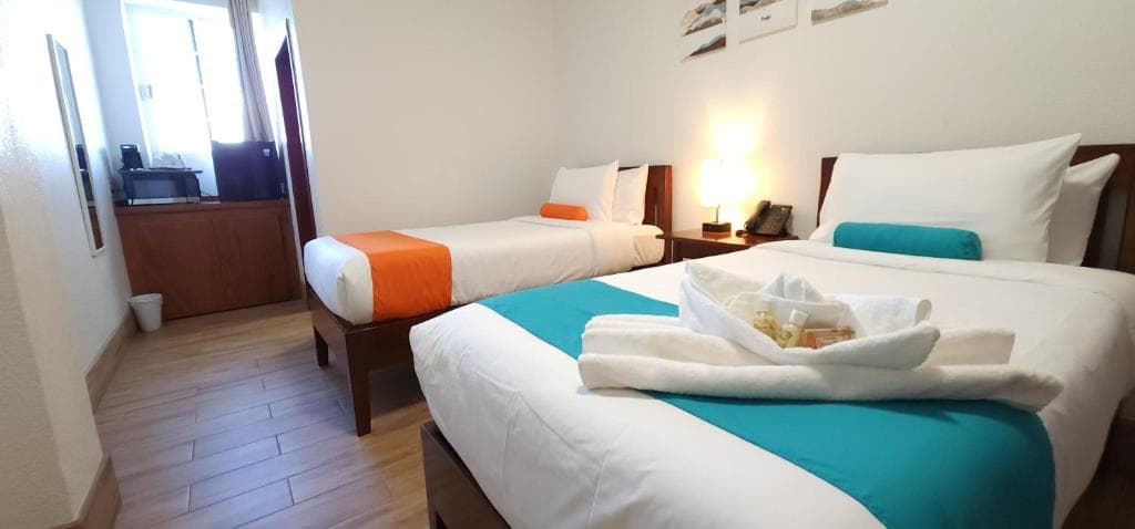 A bright hotel room featuring two beds with teal and orange accents, and a small table with refreshments.