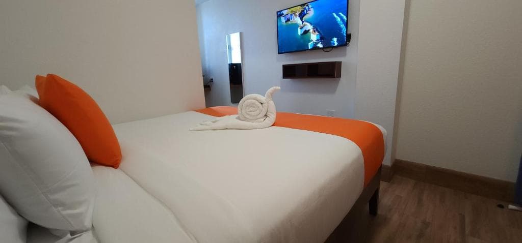 A cozy hotel room featuring a bed with orange accents, a towel arranged in a decorative shape, and a television on the wall.