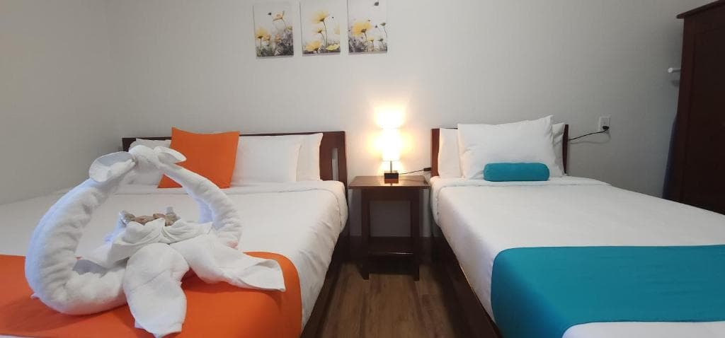 A cozy hotel room featuring two beds, one with an orange accent, decorated with a towel swan, and a bedside table with a lamp.