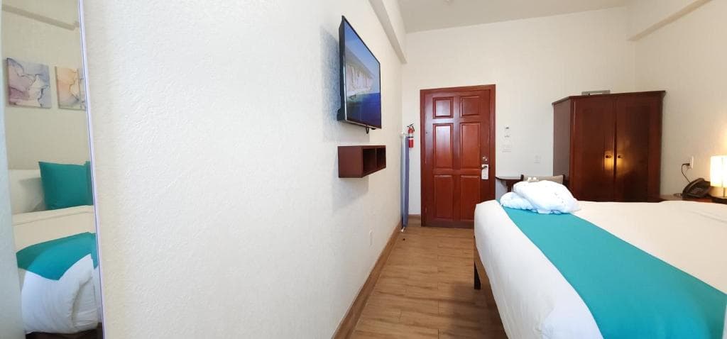 A bright hotel room featuring a double bed, a TV mounted on the wall, and a wooden wardrobe.