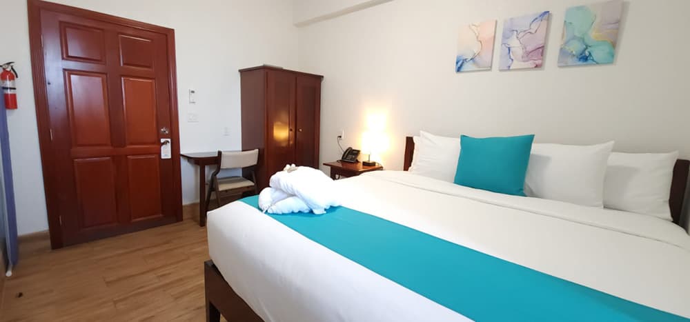 A cozy hotel room featuring a bed with turquoise accents, a wooden wardrobe, and a desk near a door.