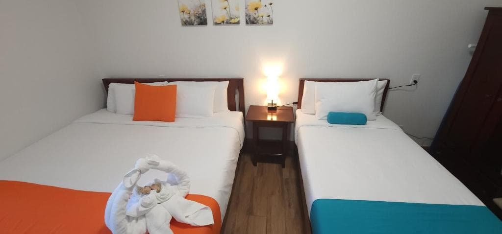 A cozy hotel room with two beds, one featuring an orange accent and the other a turquoise towel, beside a nightstand with a lamp.